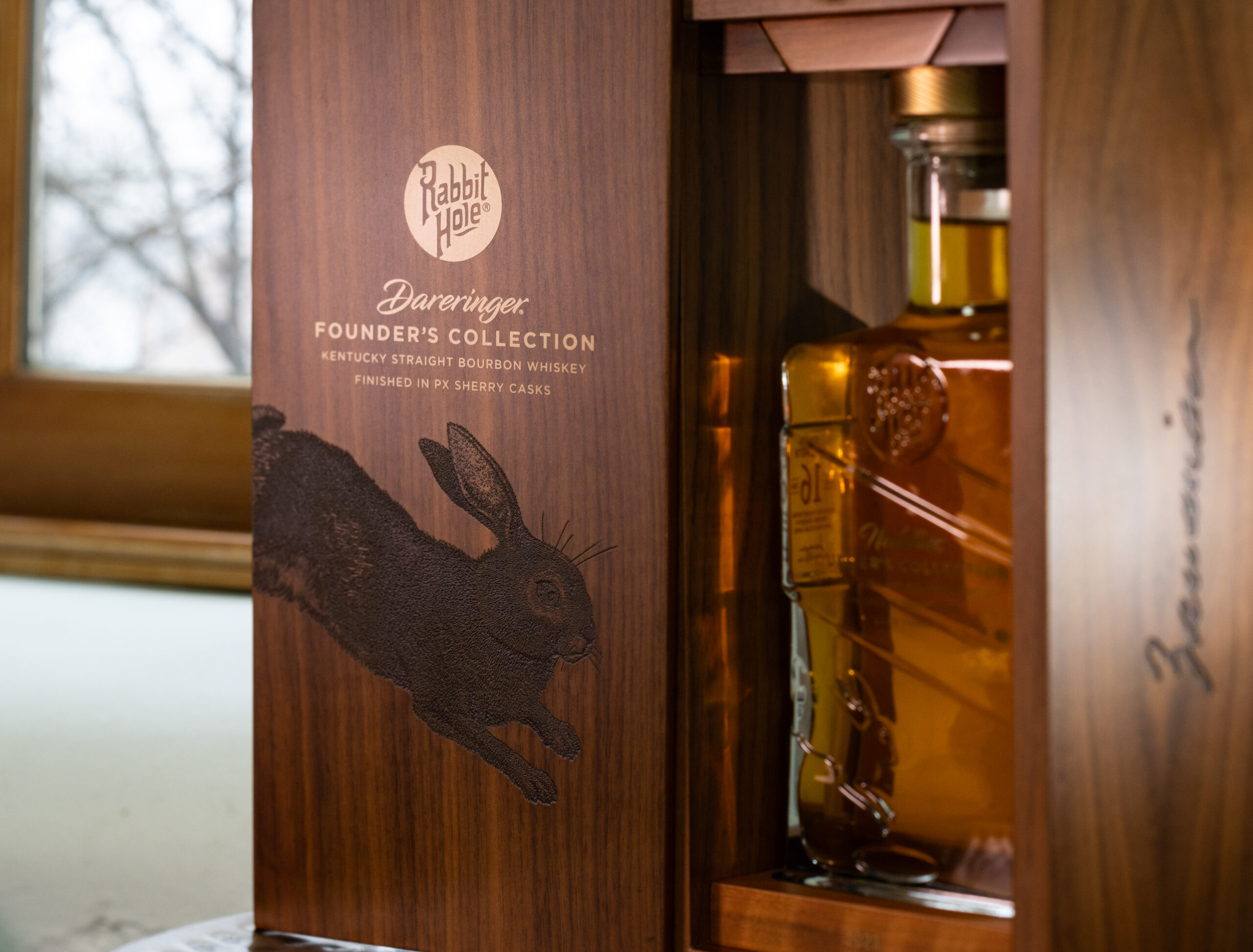brand packaging for Rabbit Hole: wooden box with rabbit design on the outside and Rabbit Hole logo. Glass bottle of alcohol inside. Text on the box says "Dareringer. Founder's Collection; Kentucky Straight Bourbon Whiskey, Finished in PX Sherry Casks."